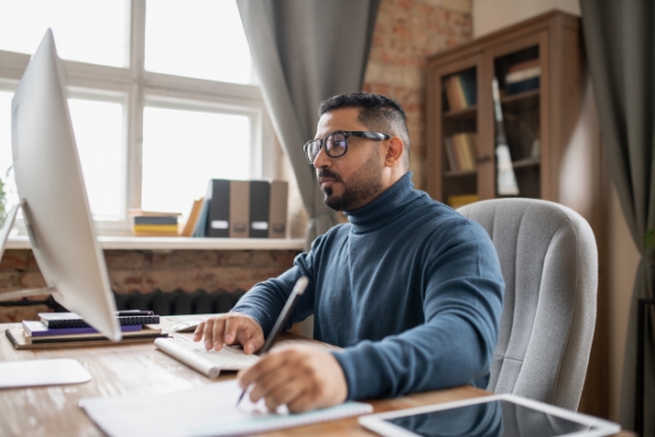 man pressing key and looking at computer screen while making notes during online lecture